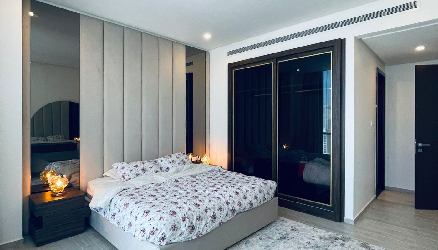 Modern bedroom with a floral-patterned bed, mirrored wall, large rug, wood flooring, bedside tables with lamps, and dark glass wardrobes.