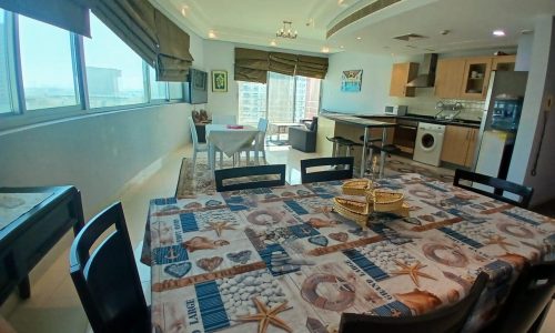 A modern kitchen and dining area with ocean-themed tablecloth, large windows, and an open-plan layout. A small dining table and chairs are visible in the background near the windows.