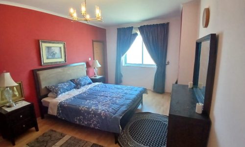 A cozy bedroom with a double bed, blue bedding, two bedside tables, lamps, a dresser with a mirror, a window with curtains, and a chandelier. The walls are red and white, with framed art on the red wall.