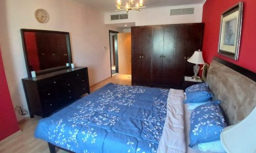 A bedroom with a blue floral bedspread, dark wood furniture, red accent wall, chandelier, and a large mirror above a dresser. A clock is mounted on the white wall. The door leads to a hallway.