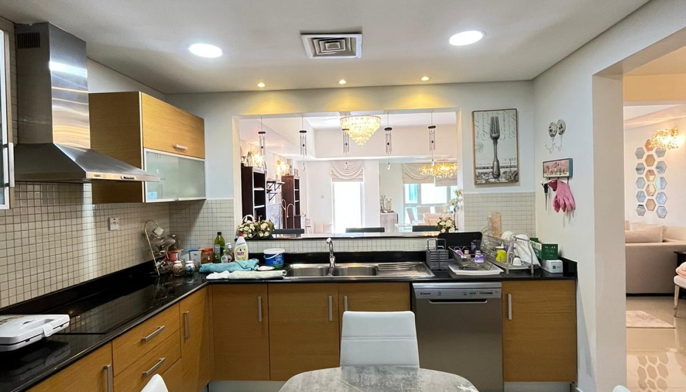 Modern kitchen with wooden cabinets, black countertop, stainless steel appliances, and a gray dining table with white chairs; view into a brightly lit living area.