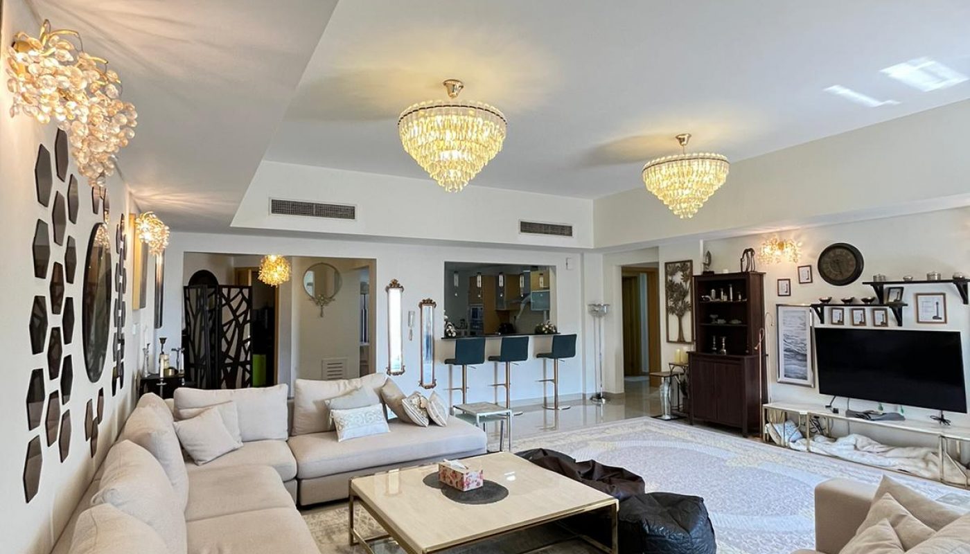 Spacious living room with beige sectional sofa, large coffee table, modern chandeliers, wall decor, a TV, and open view to the dining area and kitchen.