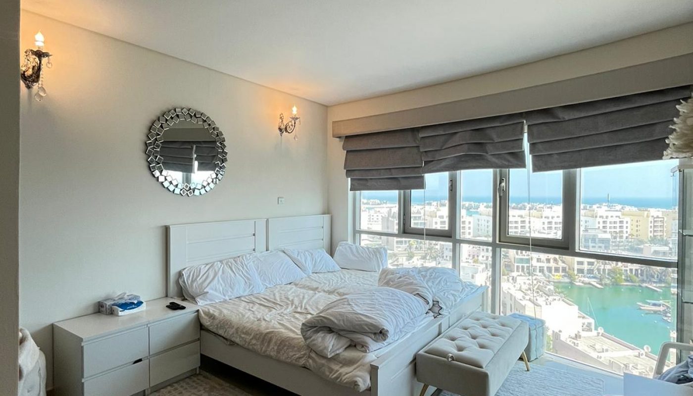 A modern bedroom with an unmade bed, two nightstands, a round mirror on the wall, large windows with gray blinds, and a view of a marina and buildings outside.
