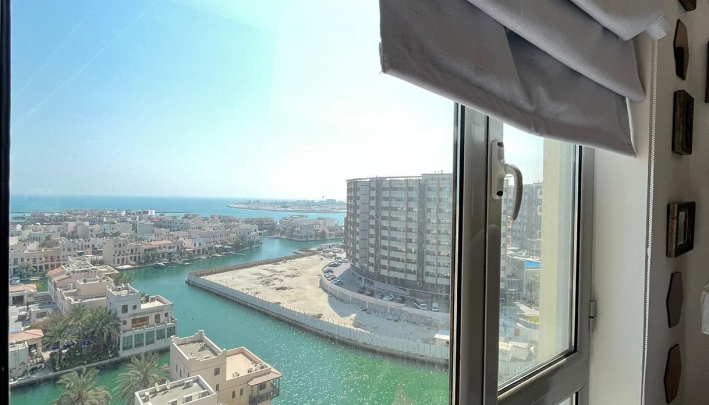 View through a window showing a canal, residential buildings, palm trees, and a clear blue sky on a sunny day.