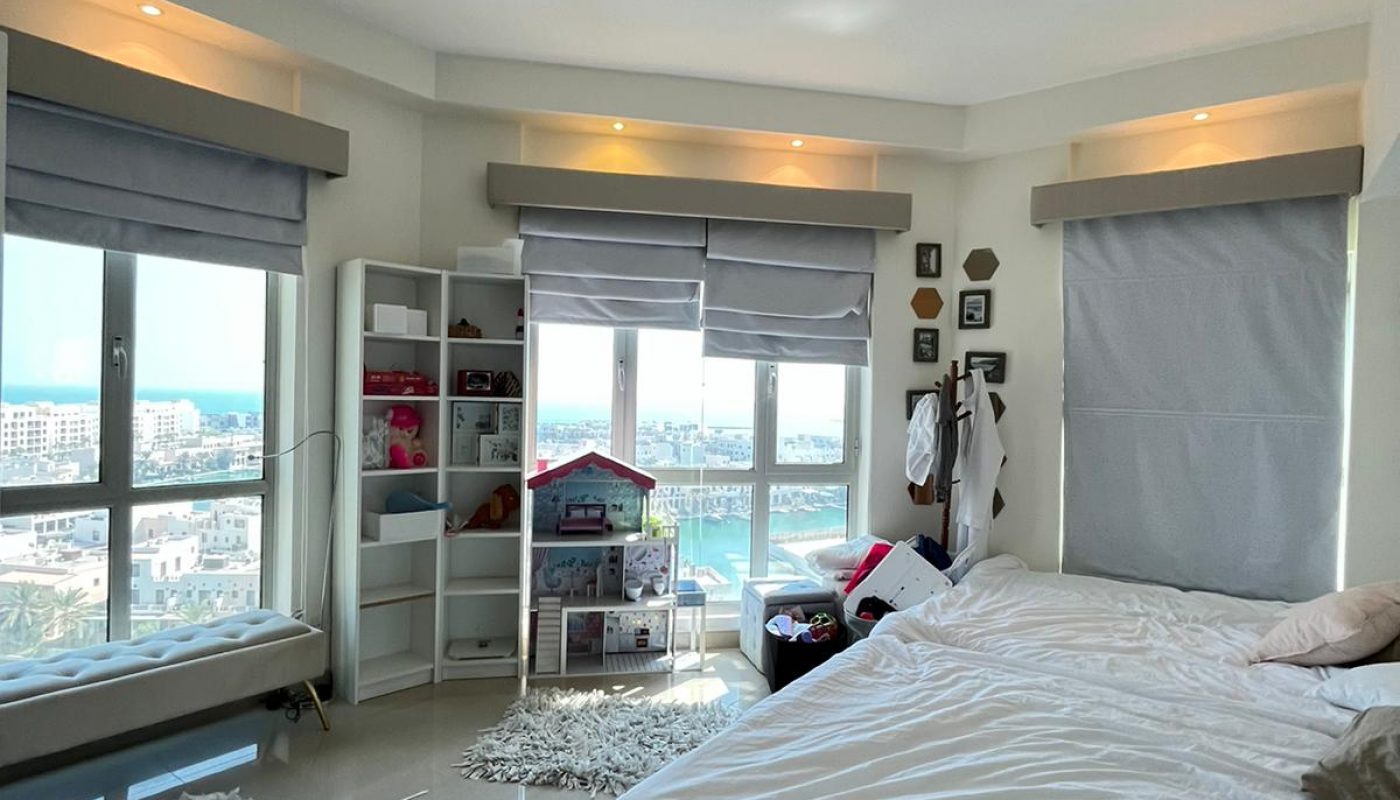 A bright bedroom with large windows, a white bed, rugs, a dollhouse, shelves, and city views outside. The room has neutral tones and modern furnishings.