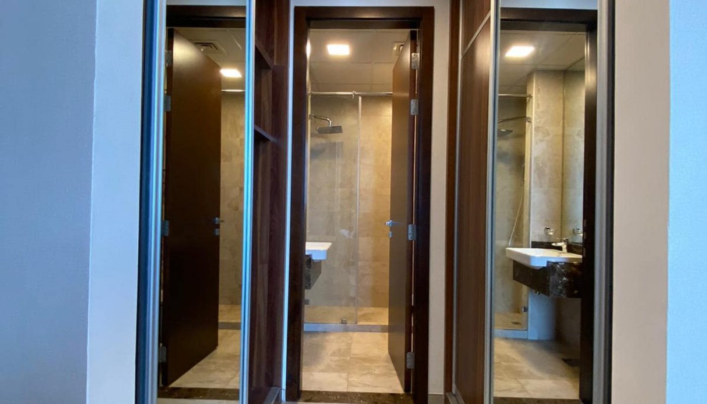 A narrow hallway with wooden flooring leads to a bathroom. Each side of the hallway has full-length mirrors, and the bathroom features a glass shower and a white sink.