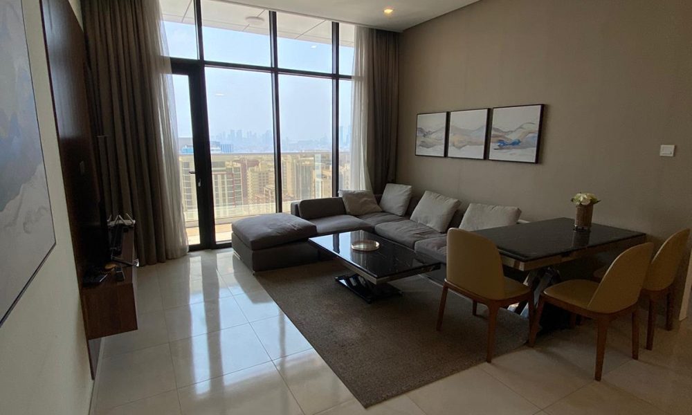 A modern living room with a large sectional sofa, a dining table with chairs, and a TV. Floor-to-ceiling windows offer a city view. Neutral tones and artwork decorate the space.