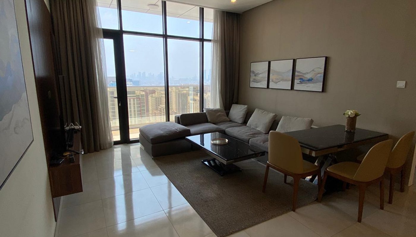 A modern living room with a large sectional sofa, a dining table with chairs, and a TV. Floor-to-ceiling windows offer a city view. Neutral tones and artwork decorate the space.