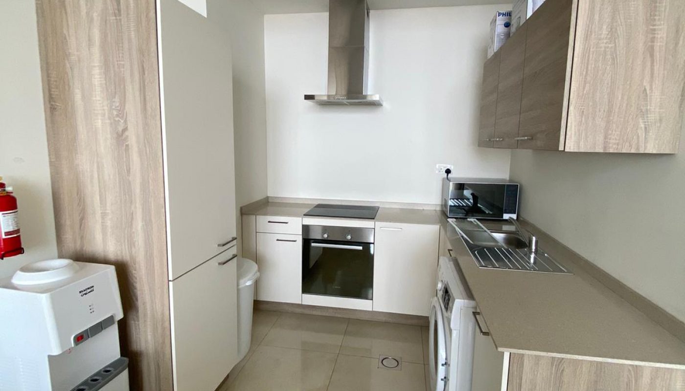 Modern kitchen with white and wood cabinets, a built-in oven, a cooktop, a washing machine, a sink, and a water dispenser. Walls and floor are light-colored.