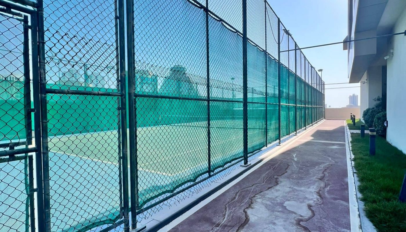 Tennis court with a green fence and weathered path, adjacent to a building under a clear sky.