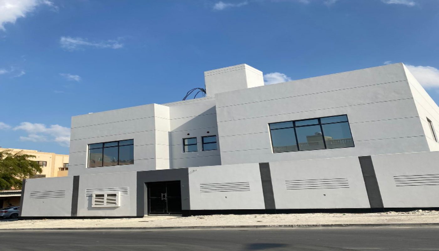 An elegant villa for sale in Hamad Town, this modern white two-story building features large windows and a flat roof, situated next to a paved road under a blue sky with scattered clouds.