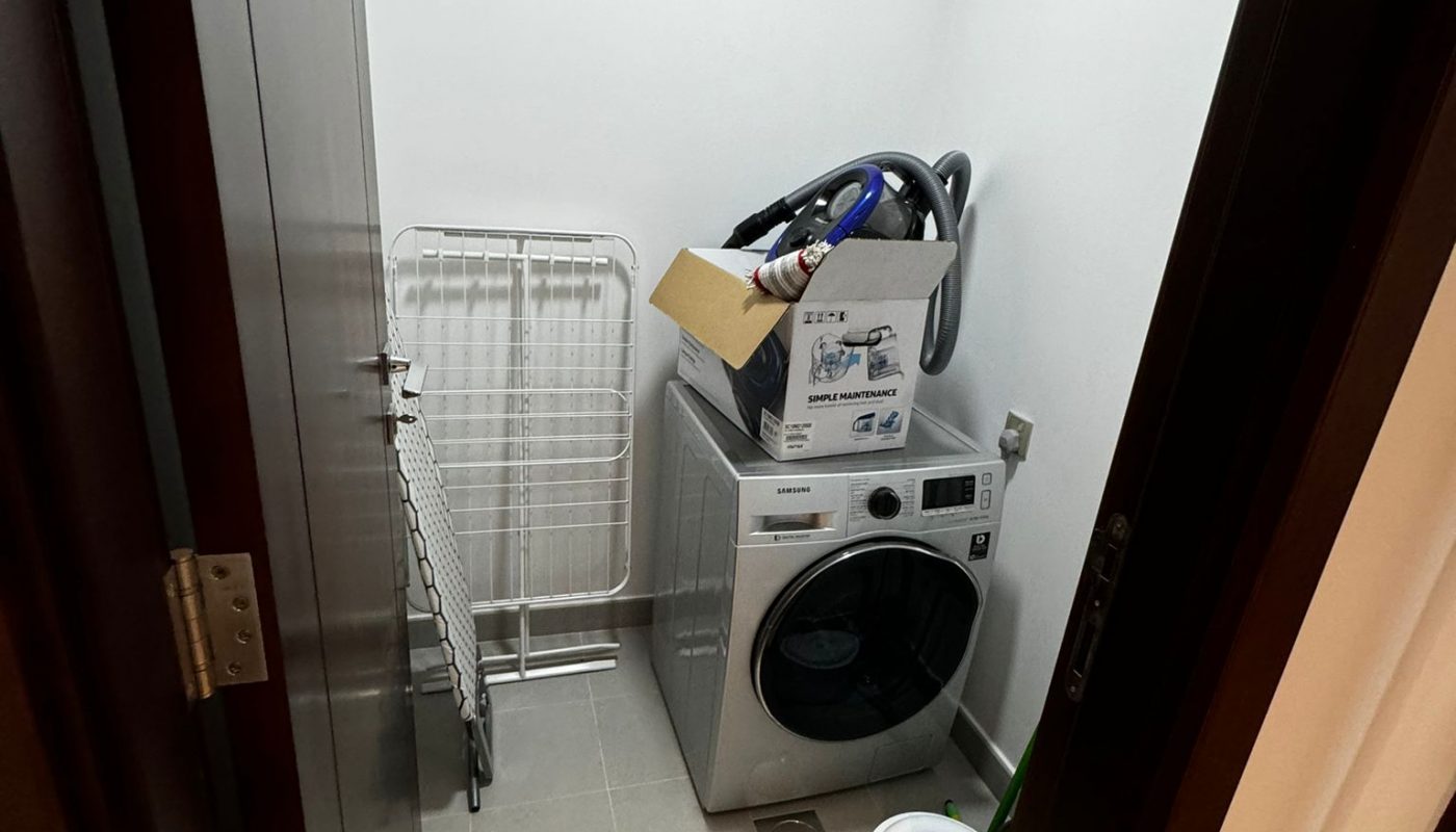 A small laundry room with a washing machine, a drying rack, a box of items, hoses, and a white bucket on the floor.