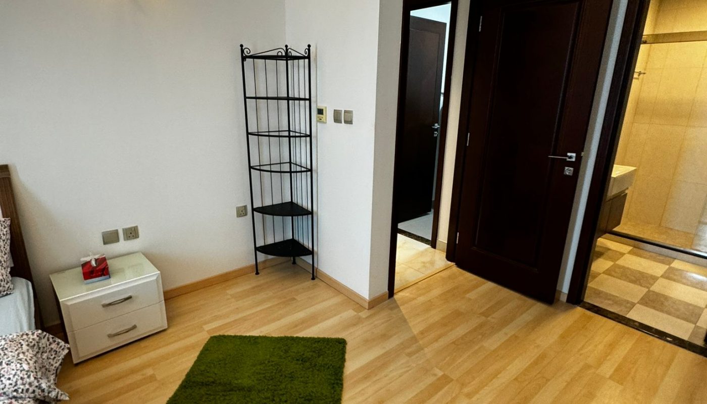 A bedroom with light wood flooring, two green rugs, a bed with patterned bedding, a white nightstand, a black metal shelf, and an open door leading to a bathroom.
