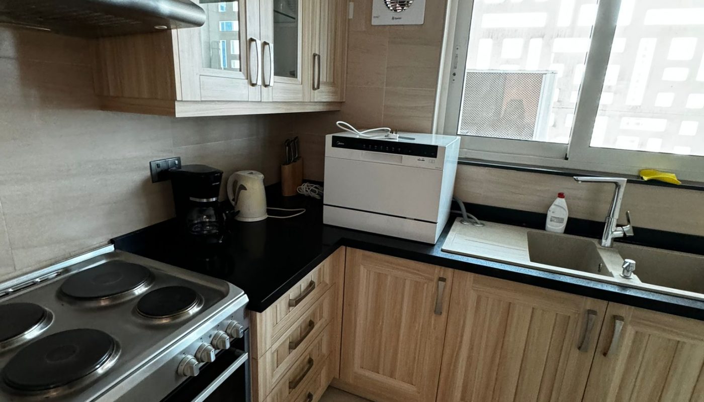 A compact kitchen with light wood cabinets, electric stove, kettle, toaster, dishwasher, sink, and a window above the countertop.