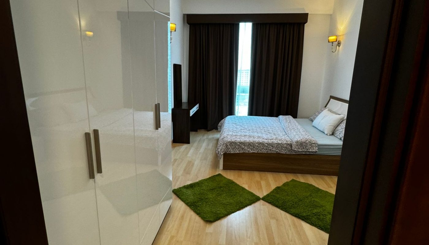 A modern bedroom with a double bed, white wardrobe, two green rugs, wood flooring, a window with dark curtains, and ceiling lights.