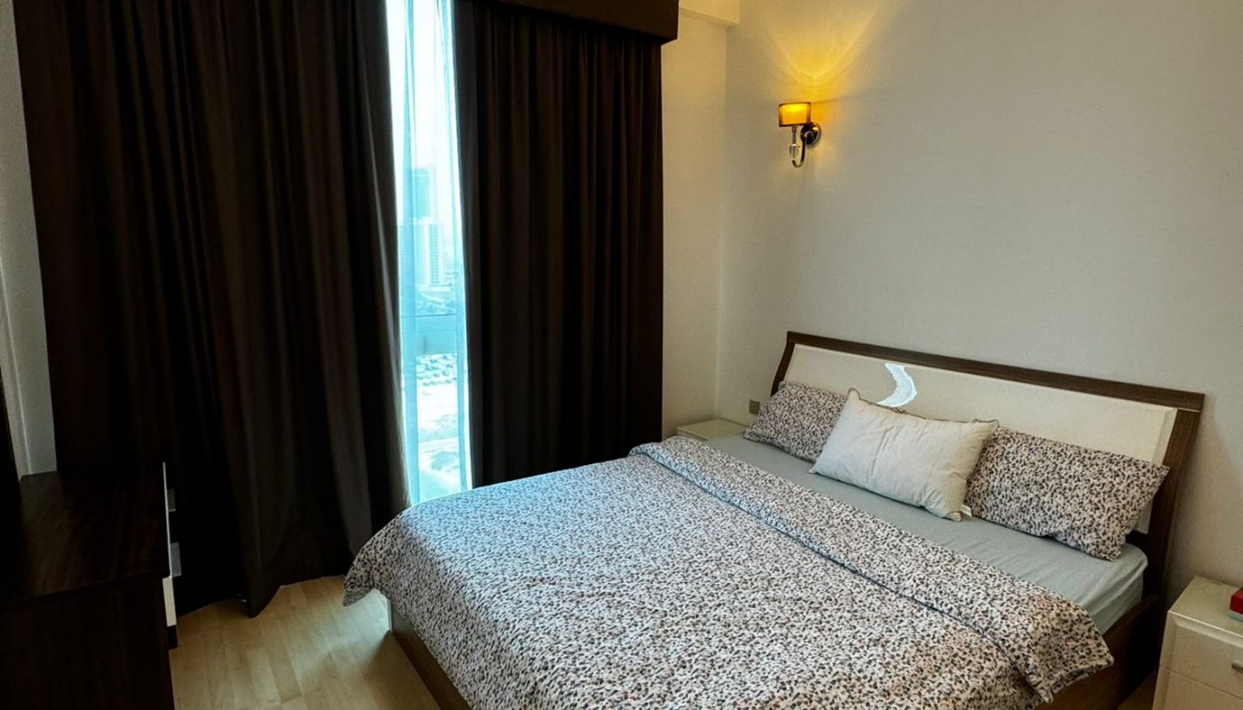 A neatly made bed with patterned bedding in a modern bedroom, featuring wooden floors, dark curtains, a chandelier, and a small nightstand.