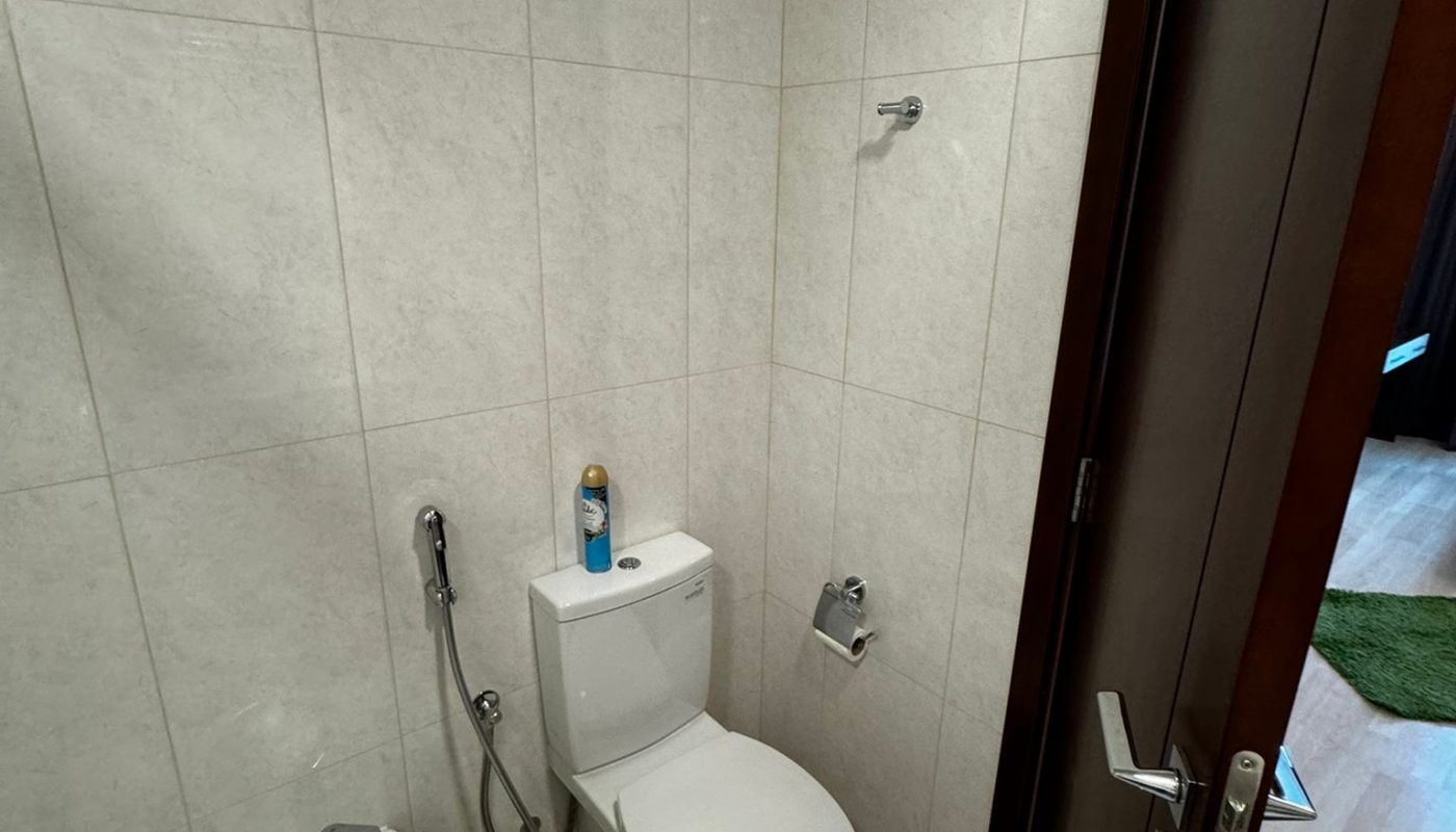 Small bathroom with a toilet, bidet, toilet paper holder, spray hose, and a bucket. Cleaning spray sits on the toilet tank. The door is partially open.