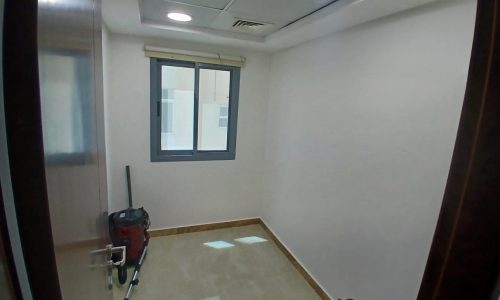 A small empty room with white walls, a tiled floor, a vacuum cleaner, and a window on one wall. Fluorescent lights are installed in the ceiling.