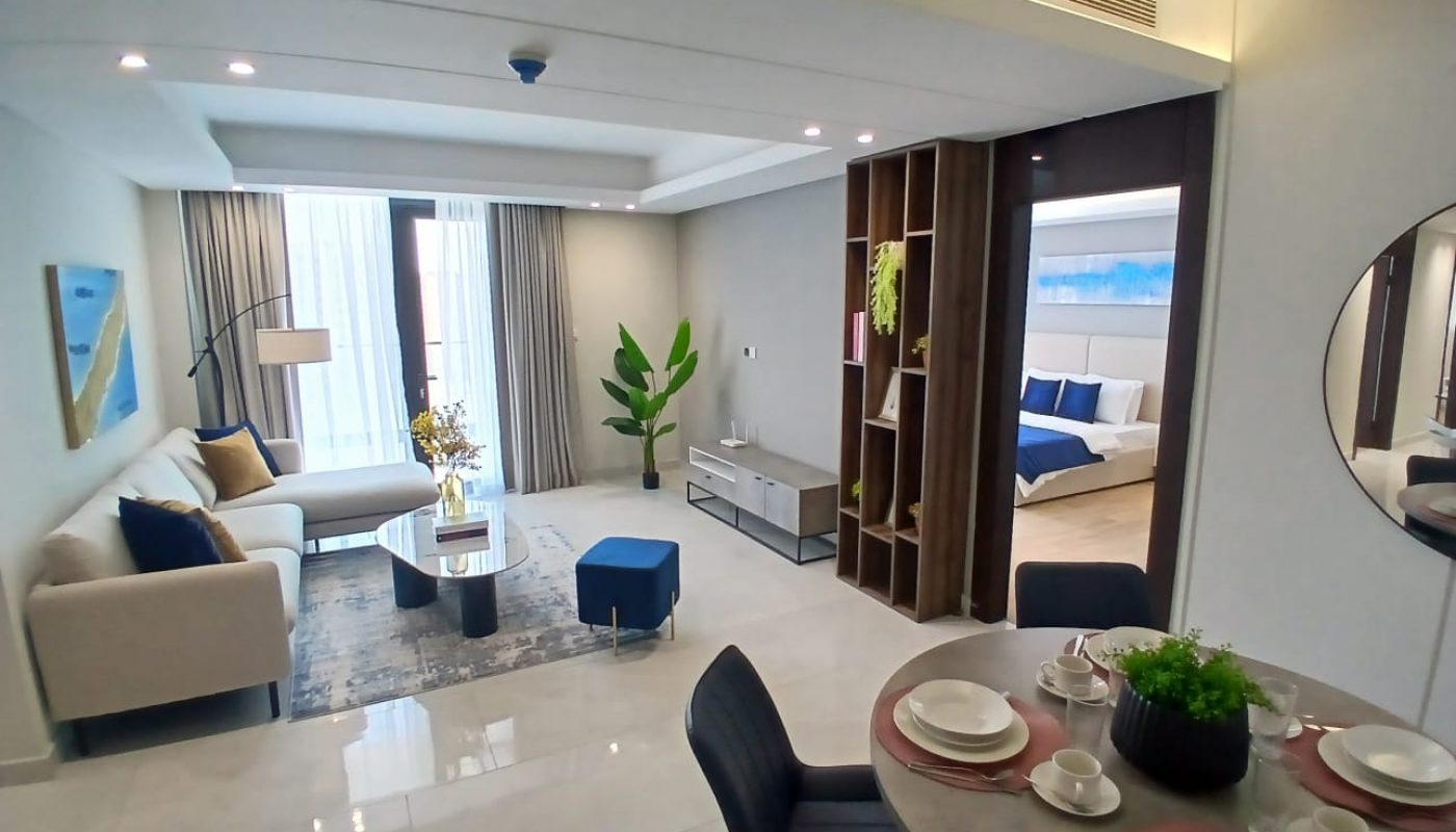 Modern open-plan living and dining area with neutral tones, a sofa, coffee table, dining set, indoor plant, and a view into a bedroom.