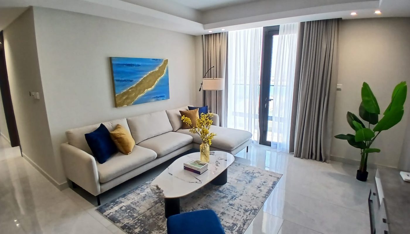 Modern living room with a beige sectional sofa, blue and gold cushions, abstract wall art, a coffee table with books and flowers, a large plant, and floor-to-ceiling windows with curtains.