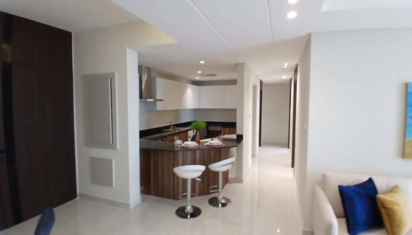 Modern kitchen and living area with white walls, wooden cabinets, two bar stools at a counter, and a sofa with blue and yellow pillows. Bright, well-lit space.