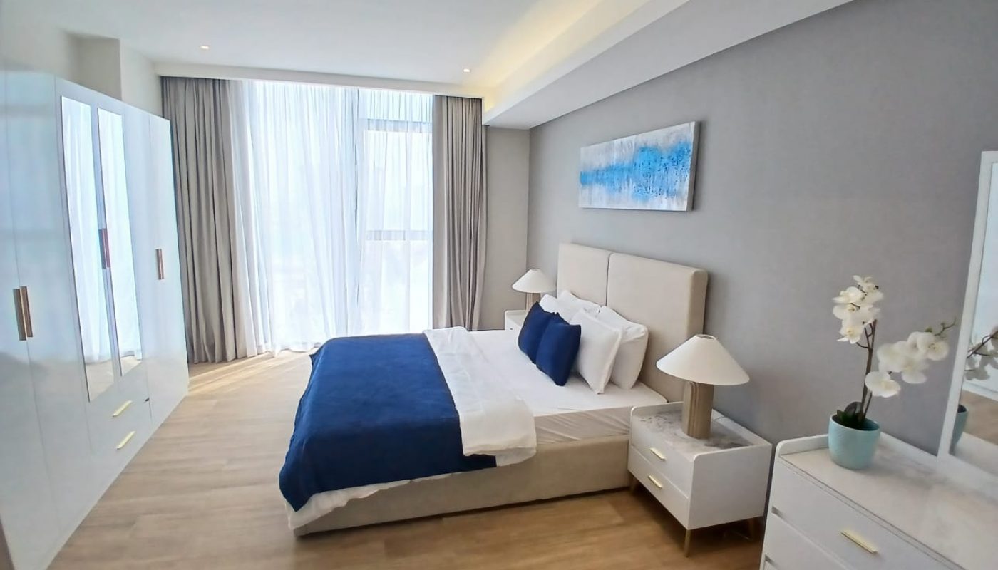 A modern bedroom with a large bed, blue and white bedding, white furniture, a wardrobe, a dresser with a potted orchid, and a window with sheer curtains.