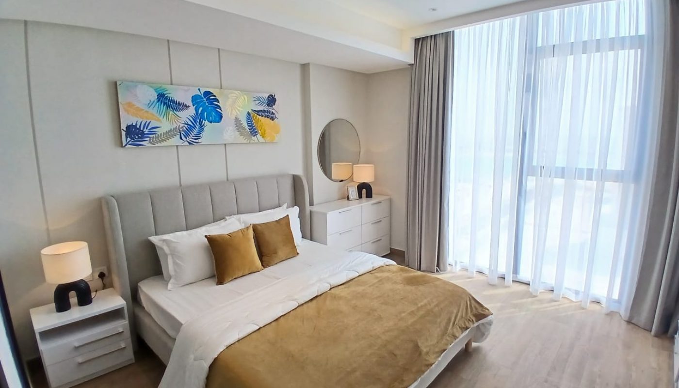 Modern bedroom with a large bed, gold bedding, two nightstands with lamps, a dresser with a round mirror, a window with sheer curtains, and a colorful leaf artwork above the headboard.