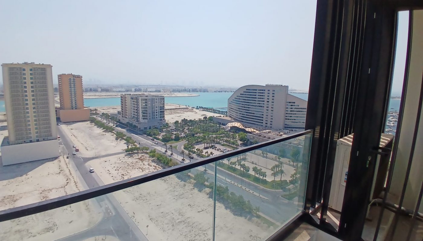 View from a high balcony overlooking a coastal cityscape with sandy areas, tall buildings, a curved structure, and the sea in the background.