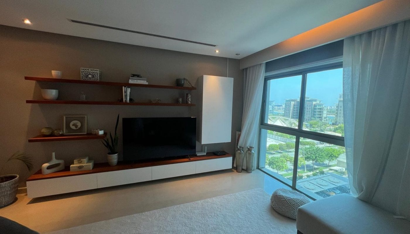Modern living room with a TV, floating shelves, and large window. Neutral tones, minimal decor, and natural light create a serene atmosphere. Curtain partially open.