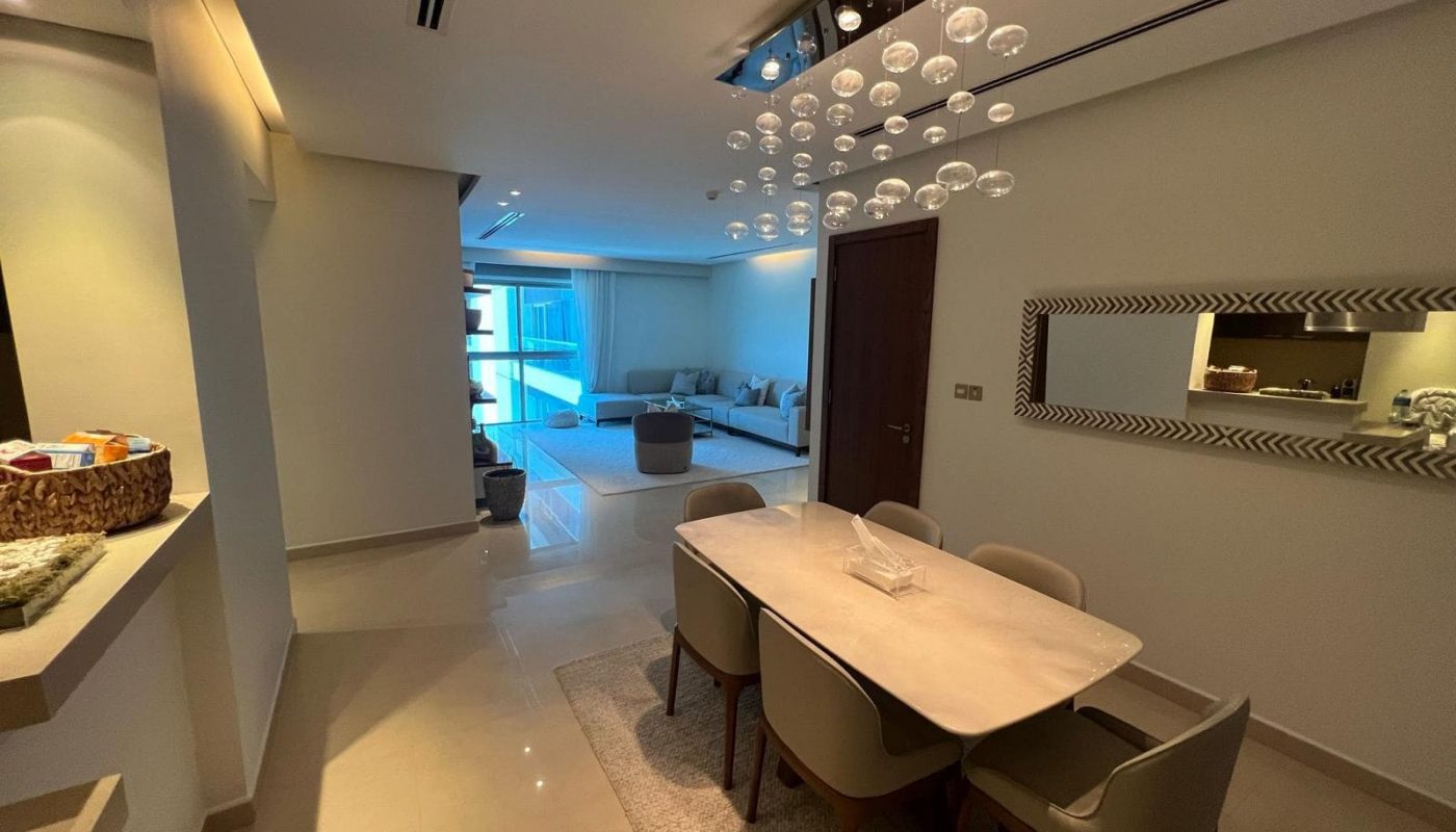 Modern dining room with a rectangular table and six chairs. Beyond is a spacious living area with a sofa and a large window. Ceiling light fixture with hanging glass spheres.