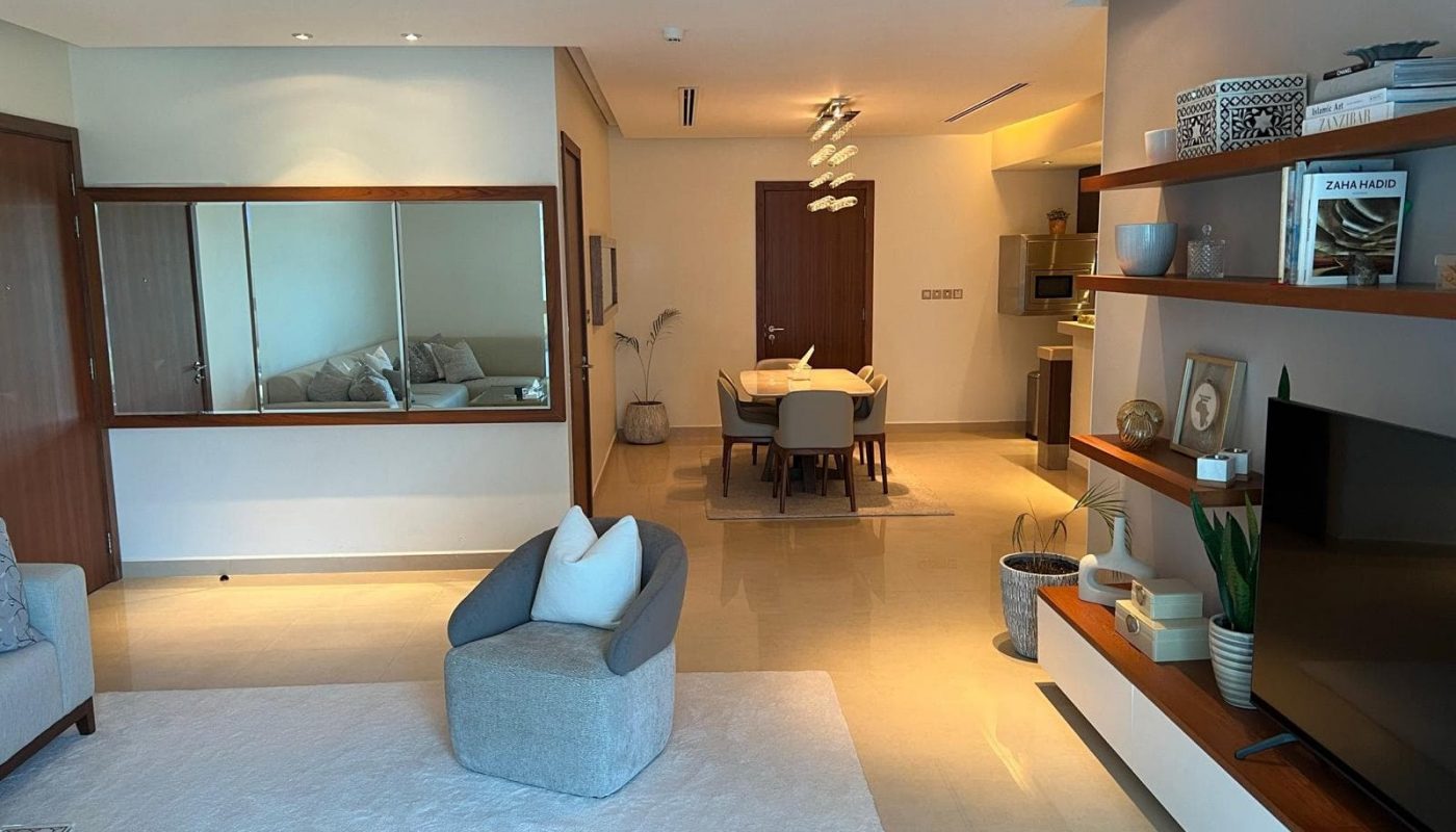 Modern living room with neutral tones, featuring a gray chair, white rug, wall mirror, and shelving unit with decor. Dining area with table and chairs visible in the background.