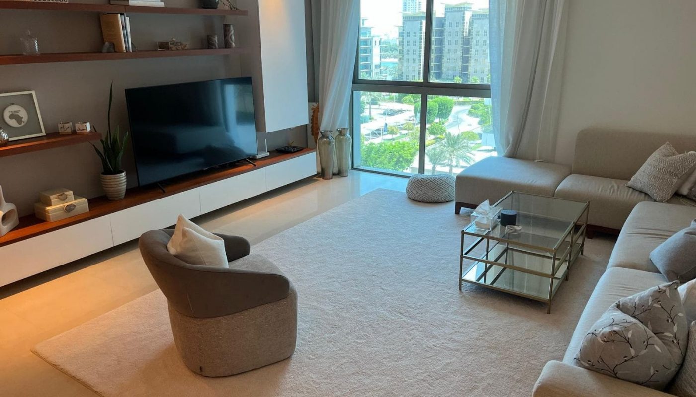 Modern living room with large windows, a white rug, gray sofas, a glass coffee table, and a wall-mounted TV on a wooden shelf. Neutral tones and decorative items create a cozy atmosphere.
