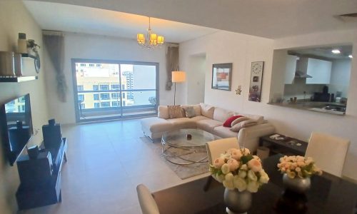 Modern living room with a sectional sofa, a glass coffee table, and a TV. Open kitchen floor plan, dining table with flower vases, large window with a city view, and contemporary lighting fixtures.