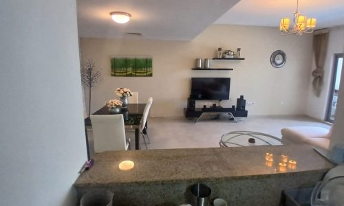 A view from the kitchen into a living room and dining area, featuring a granite countertop, dining table with chairs, wall-mounted TV, shelves, and light fixtures.