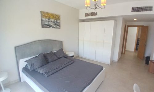 A modern bedroom featuring a gray upholstered bed with gray bedding, a white wardrobe, side tables, wall artwork, and a chandelier, with an open wooden door leading to another room.