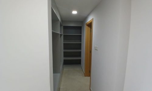 A narrow hallway with white walls, tiled floor, built-in shelving on the left, and a door on the right. A small light fixture illuminates the space.