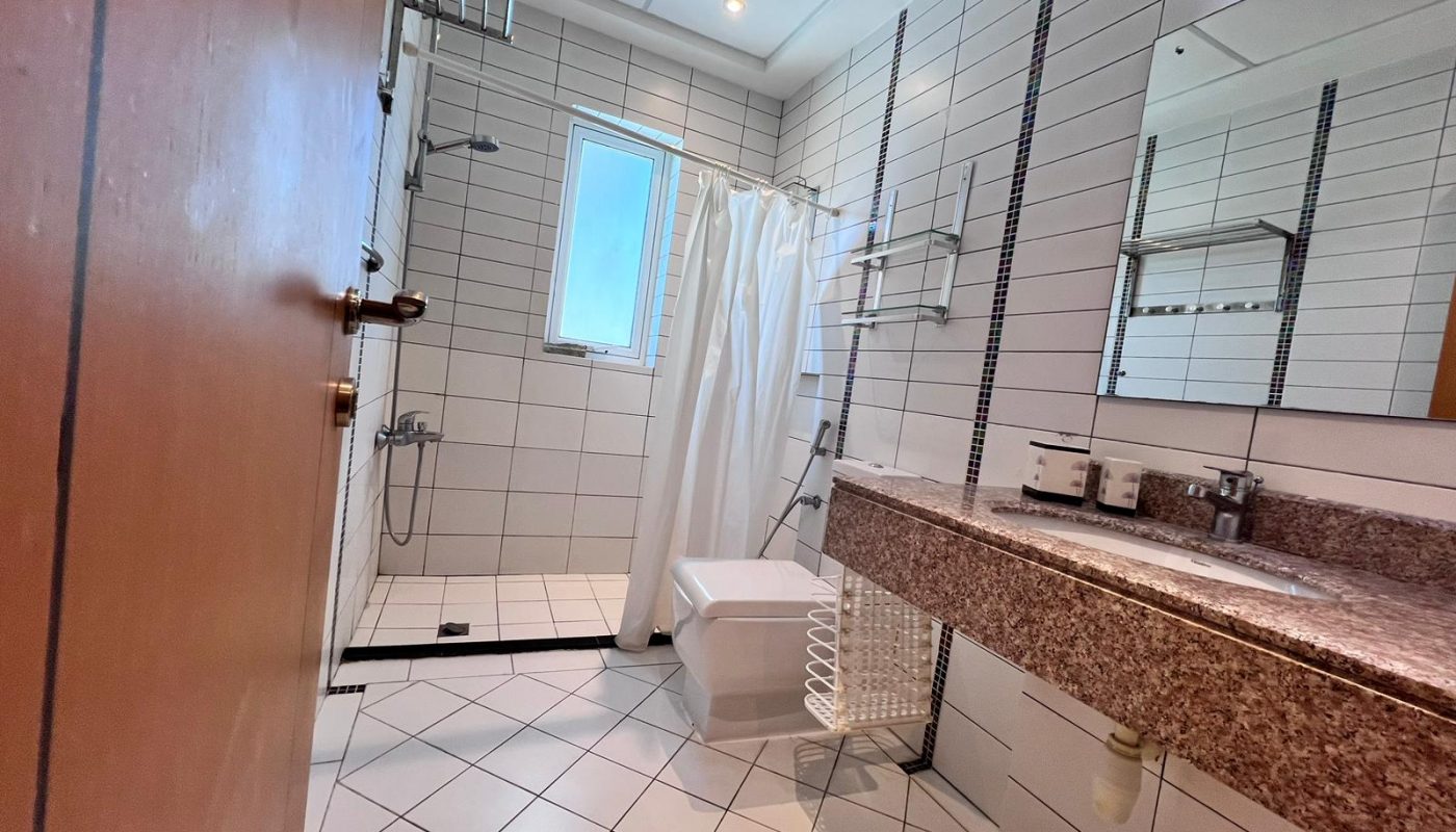 A modern bathroom with white tiled walls and floor, a shower area with a curtain, a window, a sink with a granite countertop, and a wall-mounted mirror.