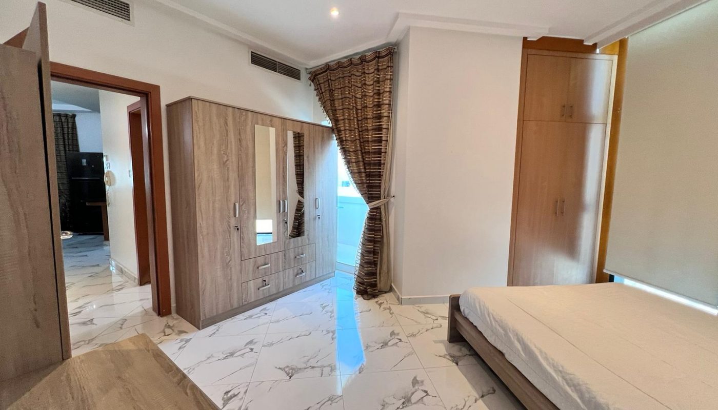 A bedroom with a bed, wooden wardrobe, chest of drawers, tiled floor, window with brown curtains, and an open door leading to another room.