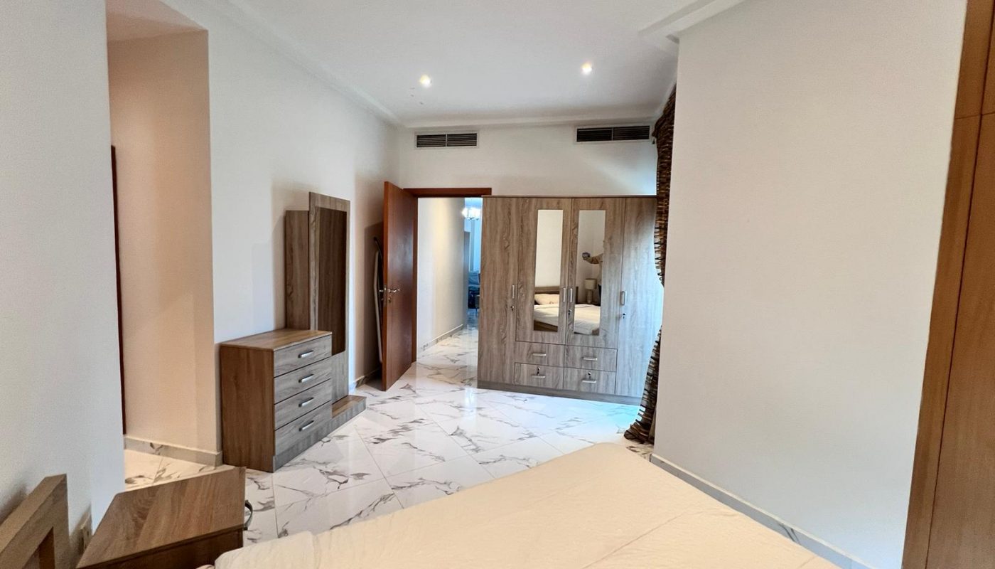 Modern bedroom with a bed, wooden wardrobes, dresser, and white marble floor; door open to a hallway with additional lighting visible.
