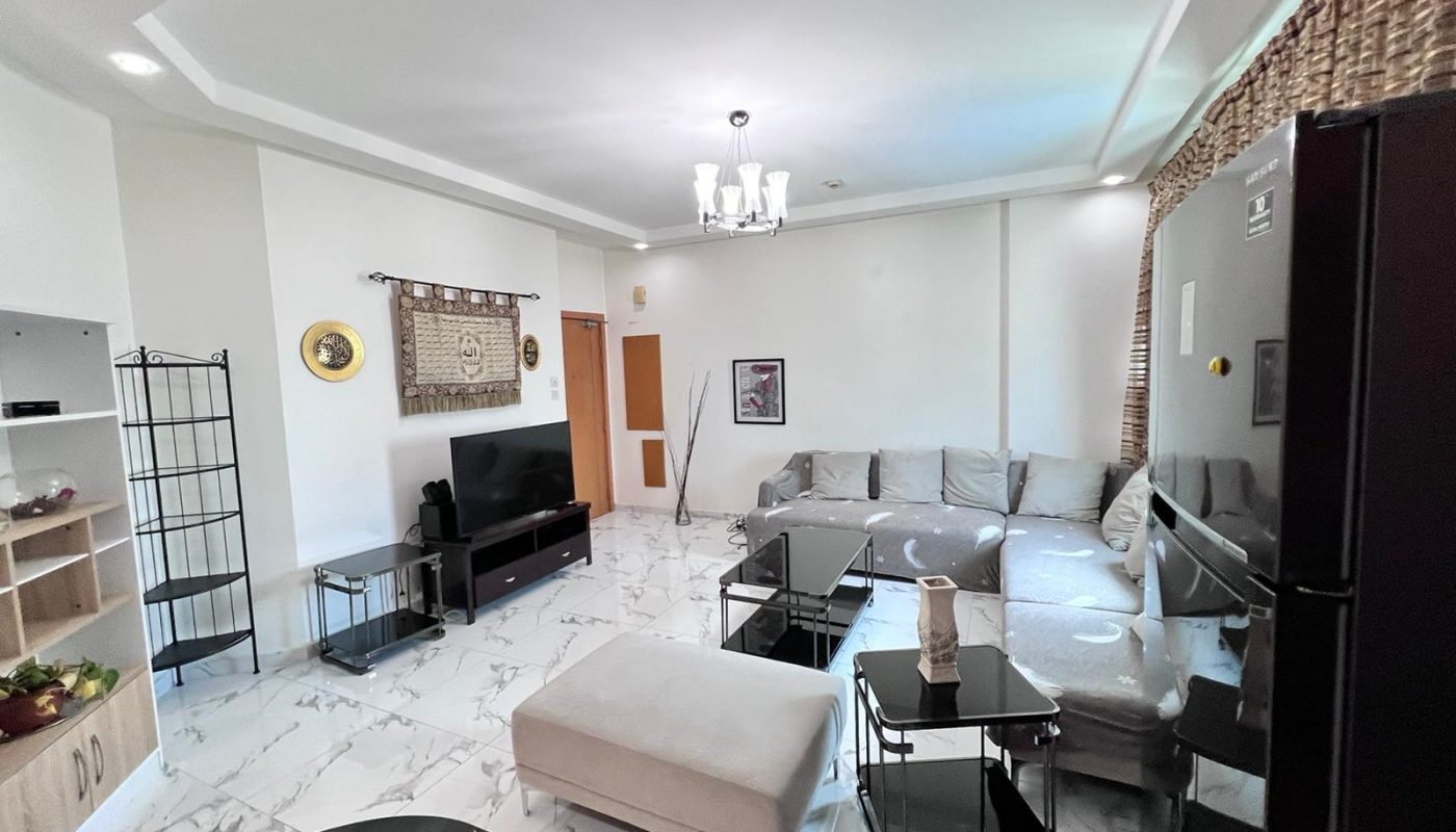 Modern living room with a gray sectional sofa, glass tables, TV, and white marble flooring, decorated with wall art and a ceiling light fixture.