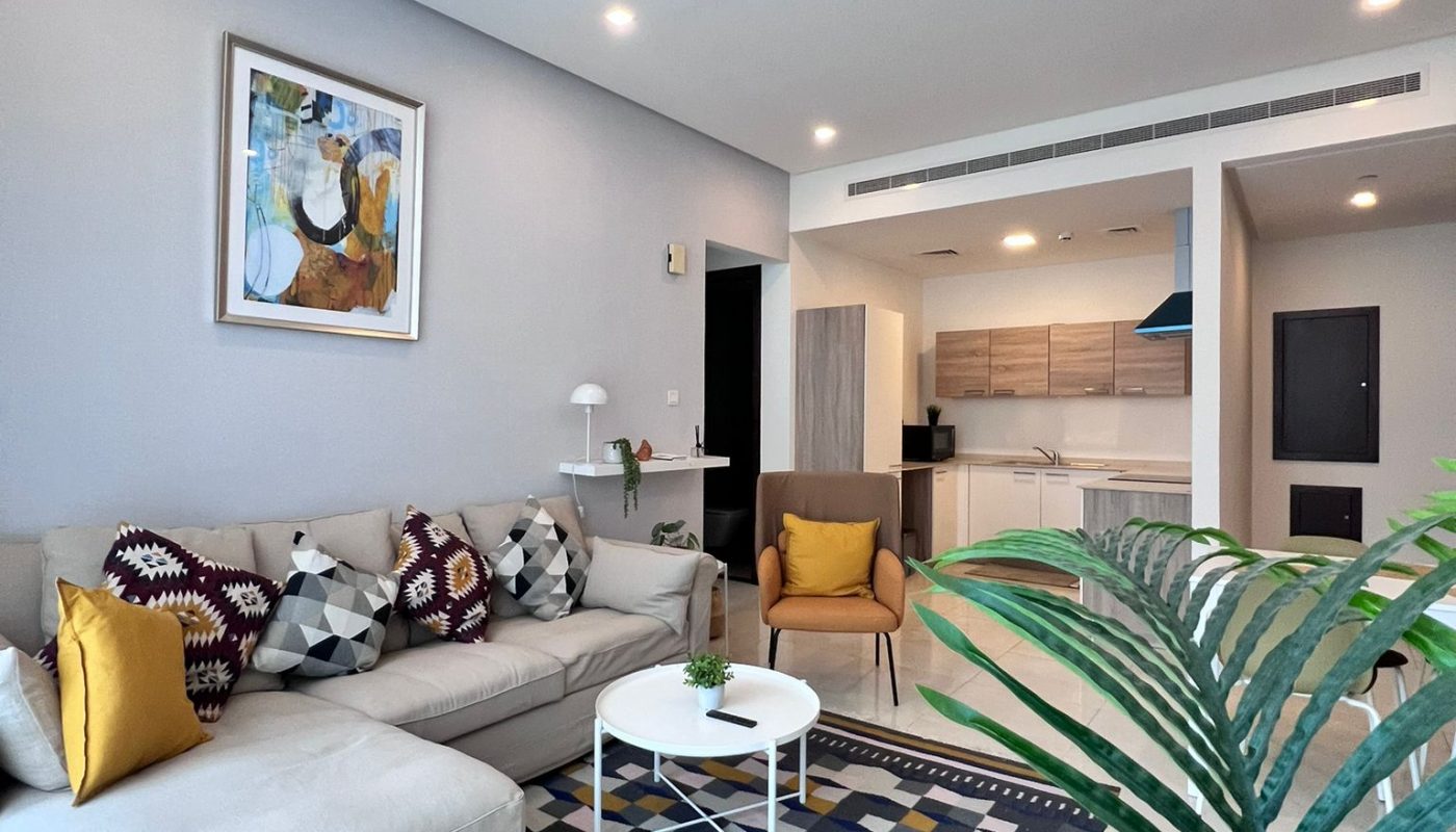 Modern living room with a sectional sofa, patterned rug, yellow armchair, wall art, and potted plant. Open kitchen is visible in the background under bright lighting.