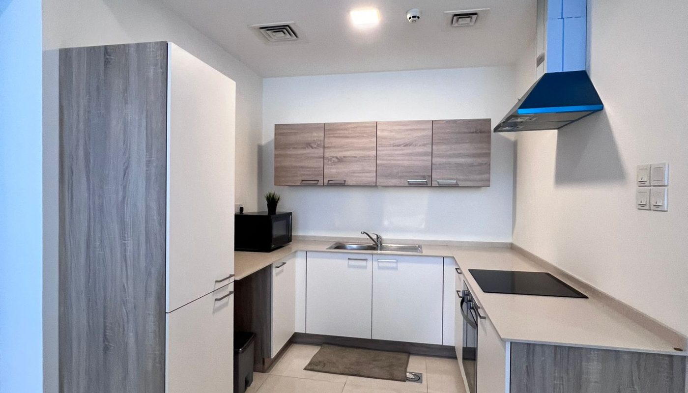 Modern L-shaped kitchen with light wood cabinets, white countertops, built-in appliances, and tiled flooring.
