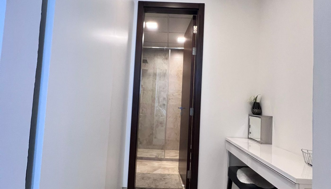 A hallway with light wood flooring leads to a bathroom with a glass shower. On the right, there is a white desk with a black stool and a small shelf.