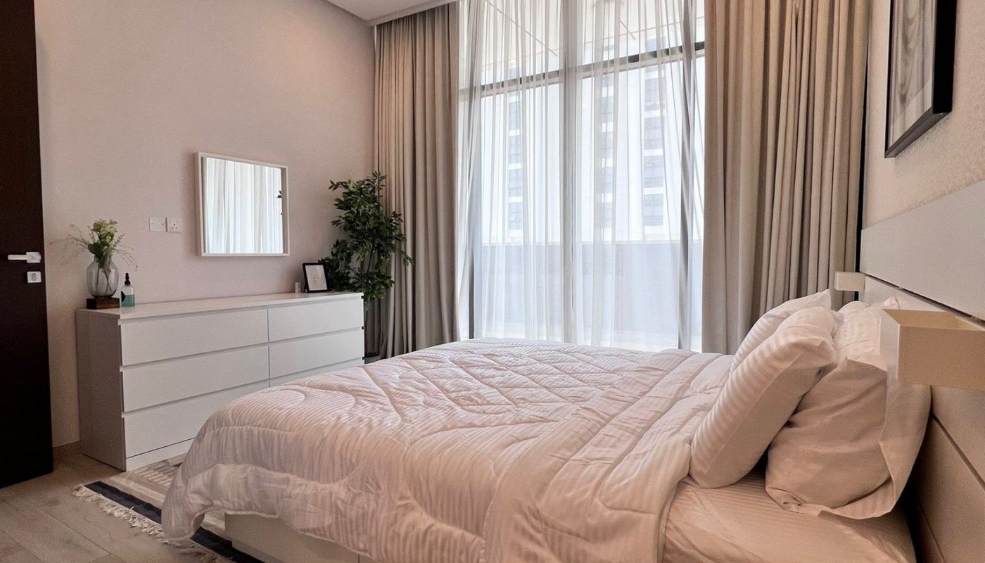 Modern bedroom with a neatly made bed, white bedding, dresser with mirror, potted plant, and large window with sheer curtains letting in daylight.