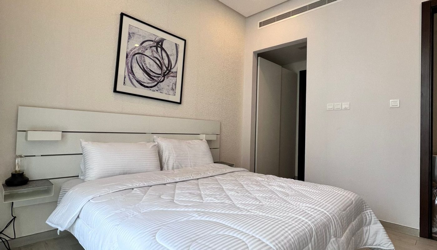 Minimalist bedroom with a white bed, abstract art above the headboard, wooden floors, and a patterned rug.