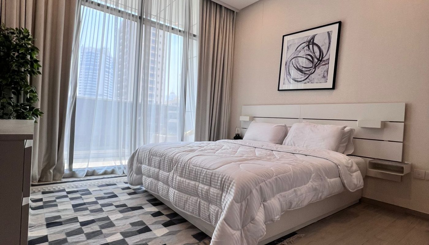 A modern bedroom with a neatly made bed, abstract art above the headboard, large windows with beige curtains, a patterned rug, light wood flooring, and a potted plant in the corner.