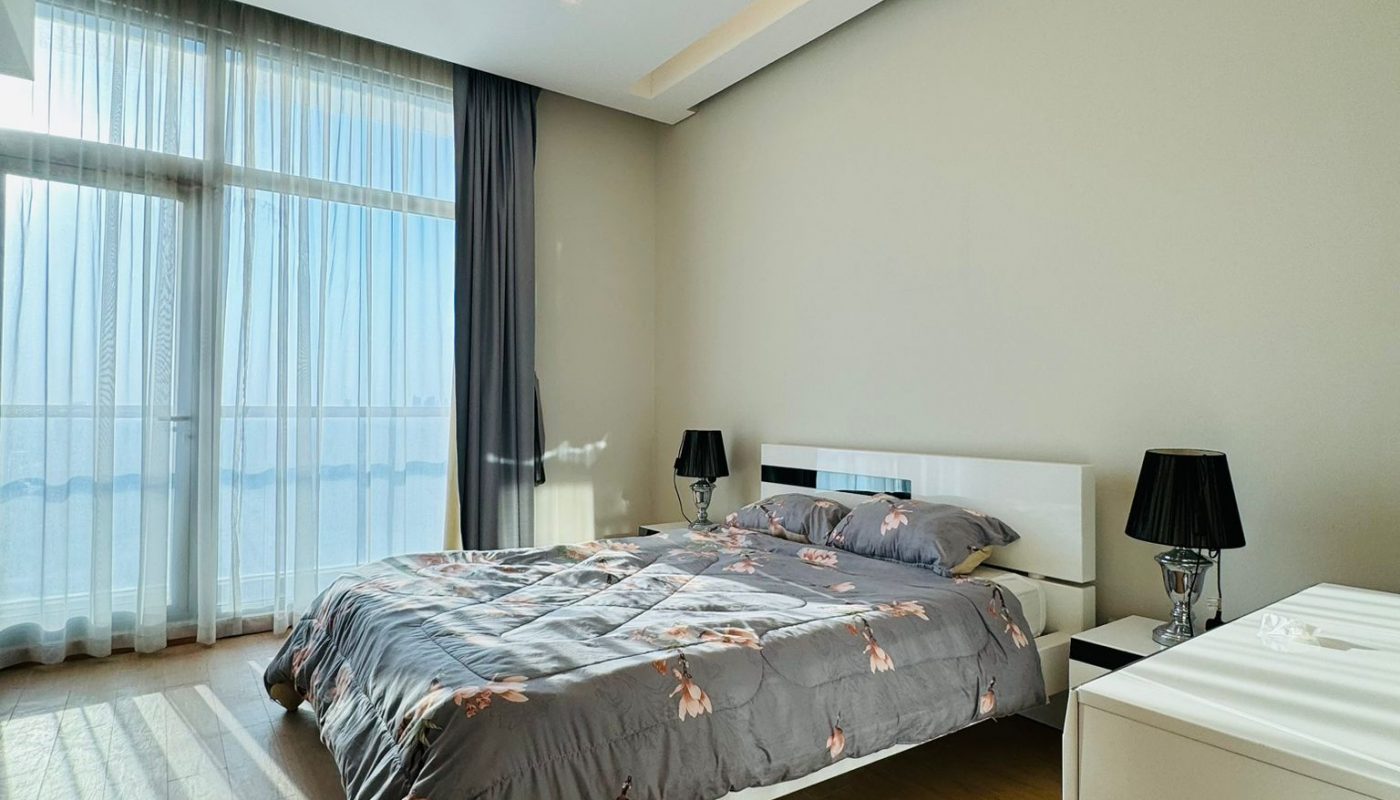 A bedroom with a queen-sized bed, floral bedding, two bedside lamps, and floor-to-ceiling windows with sheer curtains.