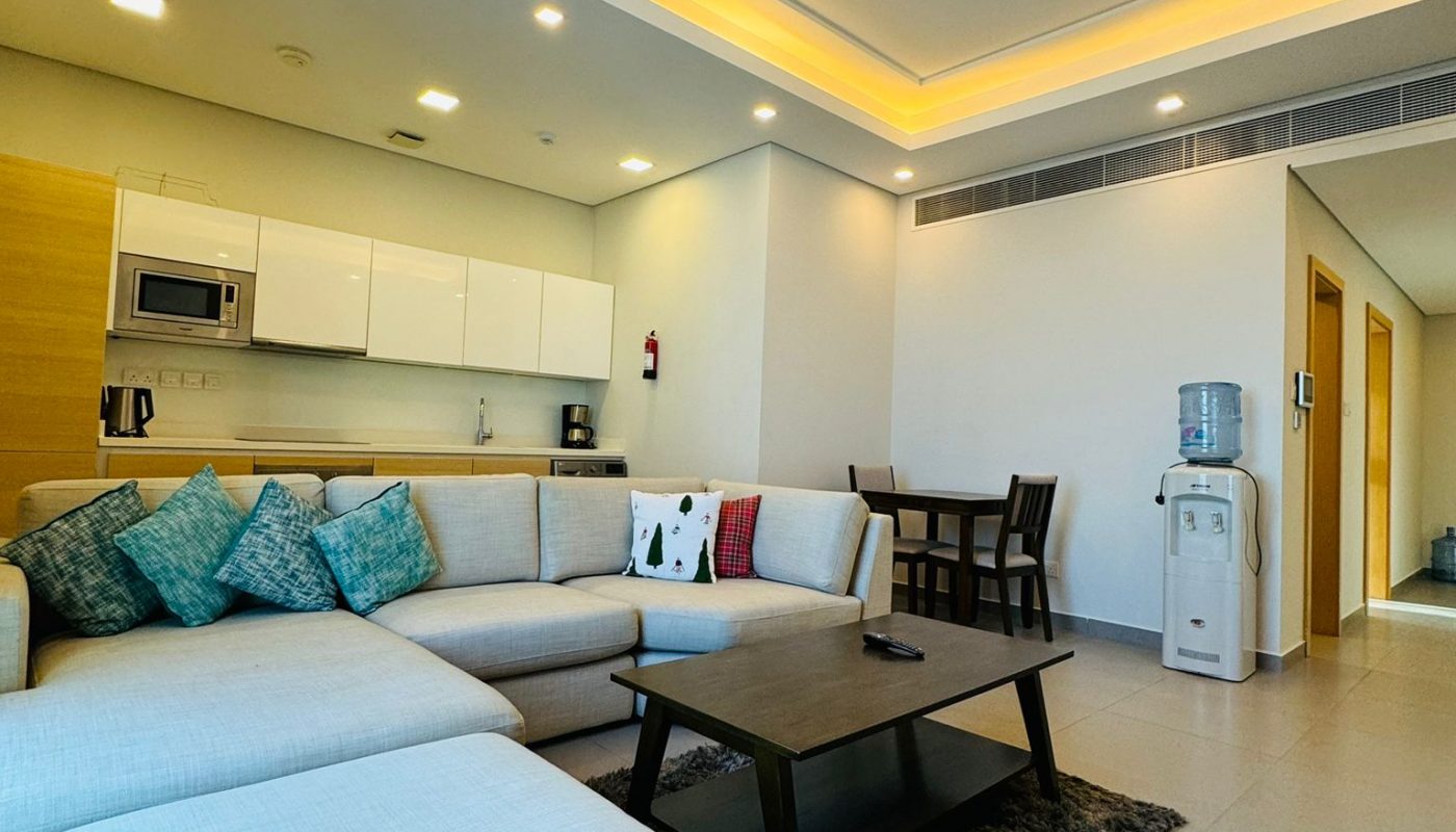 An open-concept living area with a grey sectional sofa, a coffee table, a small dining set, and a kitchenette with white cabinets.