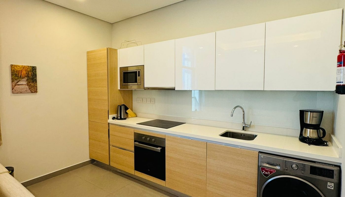 A linear kitchen with light wood lower cabinets, white upper cabinets, a built-in oven, a stainless steel washer-dryer, and a countertop with a sink and appliances.