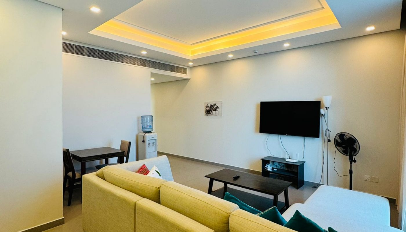 A living room featuring a light-colored sectional sofa, a coffee table, a wall-mounted television, and a small dining set in the background.